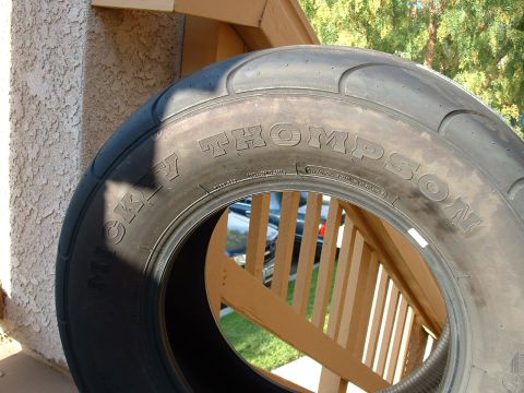 1992 Ford Crown Victoria - Close-up of an unmounted Mickey Thompson drag slick leaning against a stucco wall and wooden porch railing, with raised white sidewall lett…