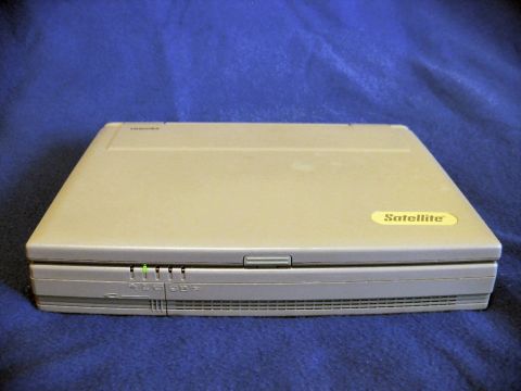 1985 Ford LTD Crown Victoria - A closed vintage beige Toshiba Satellite laptop photographed on a blue cloth backdrop, with the yellow "Satellite" badge on the lid corner…