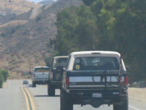 1990 Ford Bronco - Two pickups, the nearer a black Ford with a tire carrier, travel down a rural two-lane road through dry brush-covered hills.