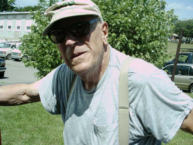 Older man in a ball cap and sunglasses wearing a gray tee at an outdoor car gathering