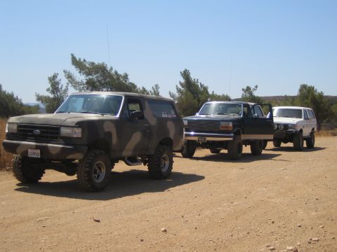 1990 Ford Bronco - Three lifted Ford trucks, led by a camouflage-painted Bronco, line up on a dirt trail amid pines under a clear sky.