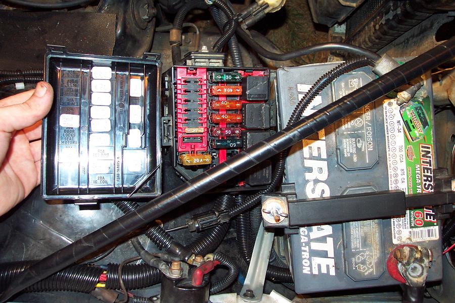 Fuse box and battery in a vehicle engine bay. A hand holds open a fuse box cover