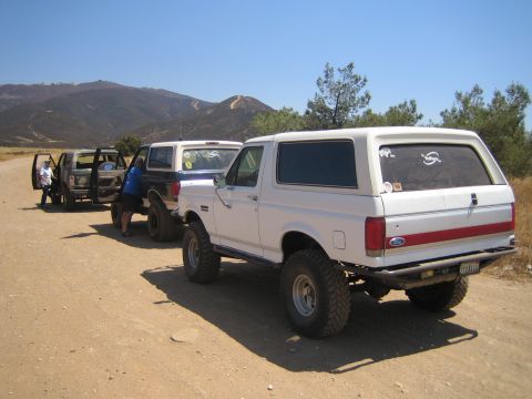 1990 Ford Bronco - Three lifted Ford Broncos, the nearest white, are parked single file on a dirt trail with people standing nearby in dry hills.