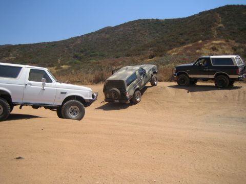 1990 Ford Bronco - Three lifted SUVs play on a dirt lot, a camouflage truck climbing a berm between a white and a black Ford Bronco amid hills.