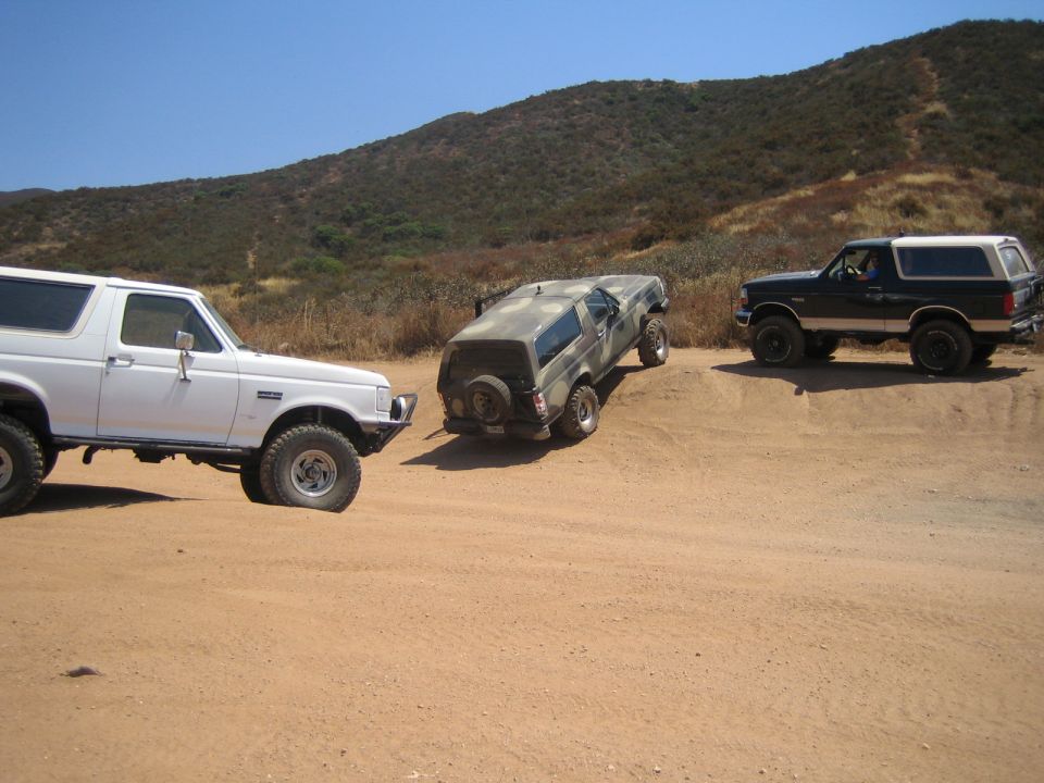 Three lifted SUVs off-road, one camo truck climbing a dirt berm between two Broncos