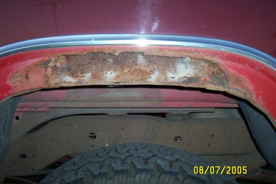 Severe rust damage on red vehicle body panel below chrome trim, with tire and undercarriage below.