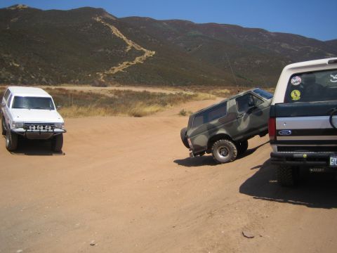 1990 Ford Bronco - white SUV and a camouflage truck maneuver on a dirt lot framed by the tailgate of a black Ford pickup in the foreground.