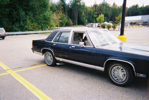 1990 Ford Crown Victoria LTD - A dark blue sedan is parked in a lot, with trees and a building in the background.