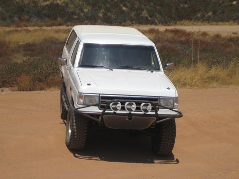 1990 Ford Bronco - white lifted Ford SUV with a camper shell and tubular front bumper carrying three off-road lights sits on a sandy dirt track.