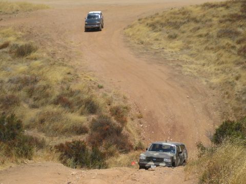 1990 Ford Bronco - Two SUVs travel along winding dirt trails cut through dry, scrub-covered rolling hills.
