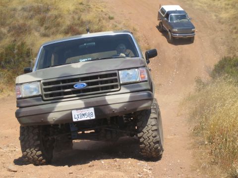 1990 Ford Bronco - camouflage-painted lifted Ford truck climbs a dirt trail head-on, a darker SUV following behind on the same path.