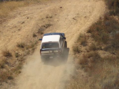 1990 Ford Bronco - Rear view of a white-roofed SUV throwing up a cloud of dust as it powers up a rutted dirt trail.