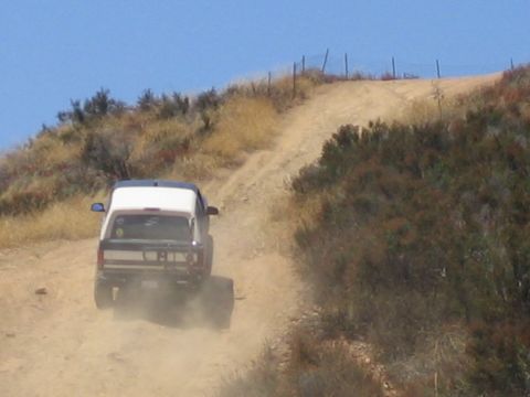 1990 Ford Bronco - white-roofed SUV grinds up a steep dusty hillside trail toward a ridgeline fence, raising dust behind it.