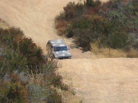 1990 Ford Bronco - camouflage-painted truck makes its way along a dirt trail flanked by dry brush and shrub-covered banks.