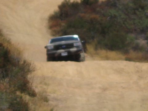 1990 Ford Bronco - Ford truck crests a dusty dirt rise head-on, its blue oval grille visible amid the brush-covered hills.