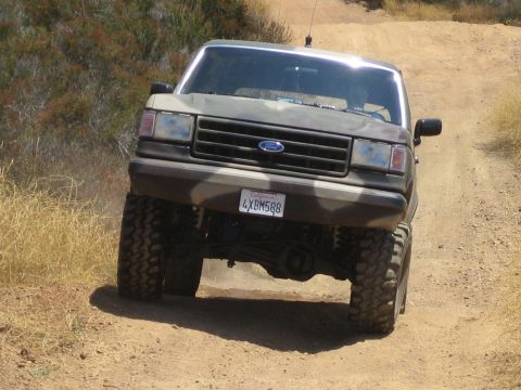 1990 Ford Bronco - camouflage-painted lifted Ford truck with a chrome billet grille and large mud tires sits on a dirt trail in dry hills.