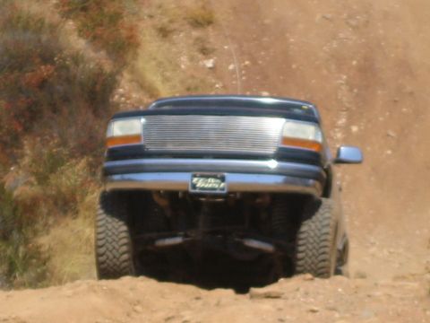 1990 Ford Bronco - black lifted Ford truck with a chrome billet grille climbs a steep dirt hill, exhaust and suspension exposed underneath.