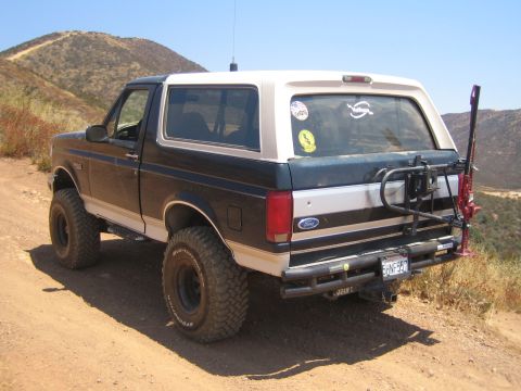 1990 Ford Bronco - lifted two-tone black and tan Ford Bronco sits on a mountain dirt trail, viewed from the rear with a swing-out tire carrier.