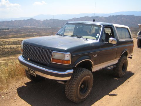 1990 Ford Bronco - black lifted Ford Bronco with a white hardtop and billet grille parked on a dirt ridge overlooking distant mountains.