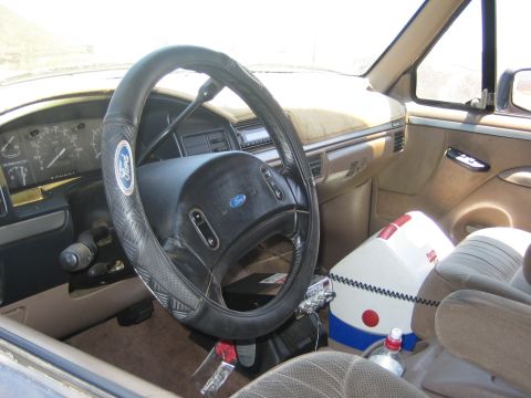 1990 Ford Bronco - Tan interior of a Ford truck with a Ford-badged steering wheel cover, gauge cluster and cloth seats, a cooler beside the seat.