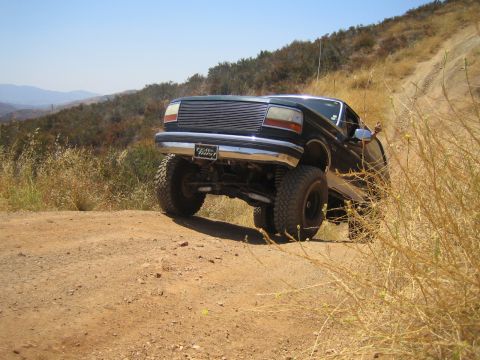 1990 Ford Bronco - black lifted Ford with a billet grille articulates over a steep dirt hill on a trail, front wheel lifted off the ground.