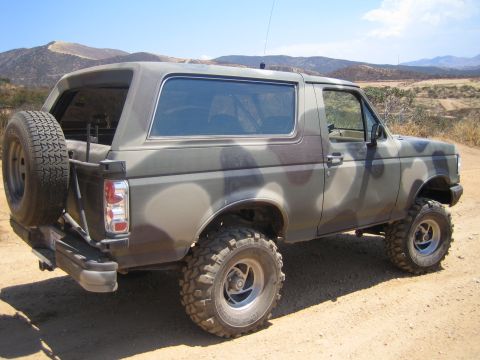1990 Ford Bronco - camouflage-painted lifted Ford Bronco with exterior spare tire sits on a dirt trail under a clear sky.