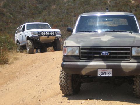 1990 Ford Bronco - Two lifted Ford trucks on a desert dirt trail, a gray one with California plate 4XBM588 in front and a white Bronco behind.