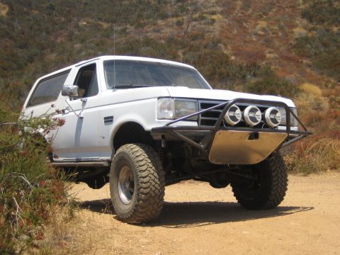 1990 Ford Bronco - white lifted Ford Bronco with a tubular front bumper, skid plate and four round off-road lights parked on a dirt trail.