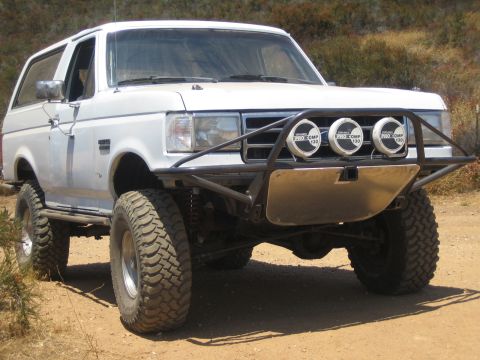 1990 Ford Bronco - white lifted Ford Bronco with a tubular front bumper and three Pro Comp round off-road lights on a sandy trail.
