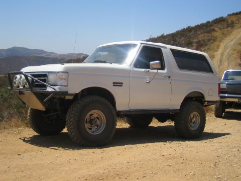 1990 Ford Bronco - white lifted Ford Bronco with a tubular front bumper and off-road lights parked on a dirt ridge beside another truck.