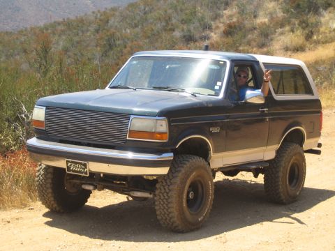 1990 Ford Bronco - black and tan lifted Ford Bronco with billet grille on a dirt trail, the driver flashing a peace sign out the window.