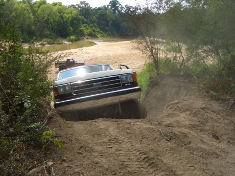 1990 Ford Bronco - A 1990 Ford F-Series truck navigates a challenging dirt path near a river, with another truck visible in the background.