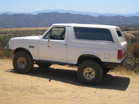 1990 Ford Bronco - white lifted Ford Bronco in side profile parked on a dirt ridge with a hazy mountain range in the background.