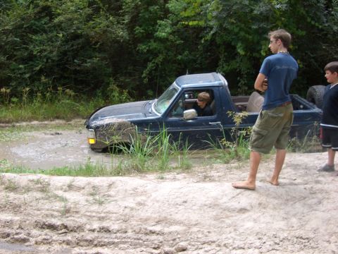 1990 Ford Bronco - A dark blue pickup truck is stuck in a muddy ditch with vegetation on the sides.