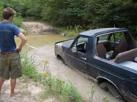 1990 Ford Bronco - A person watches as a dark blue 1990 Ford Bronco becomes stuck in a muddy off-road path.