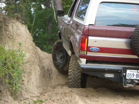 1990 Ford Bronco - A maroon and silver Ford Bronco navigates a sandy trail, showing its rear end and off-road tires.
