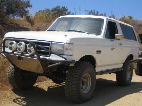 1990 Ford Bronco - white lifted Ford Bronco with a tubular front bumper and three Pro Comp lights parked against a brushy dirt embankment.