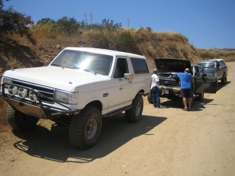 1990 Ford Bronco - white lifted Ford Bronco parked on a dirt road as two men work under the raised hood of a second truck behind it.