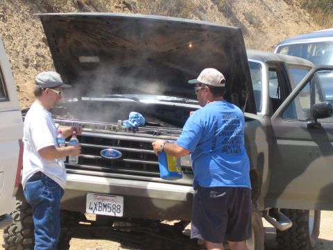 1990 Ford Bronco - Two men tend a steaming Ford truck engine with the hood up on a dirt trail, California plate 4XBM588 visible below the grille.