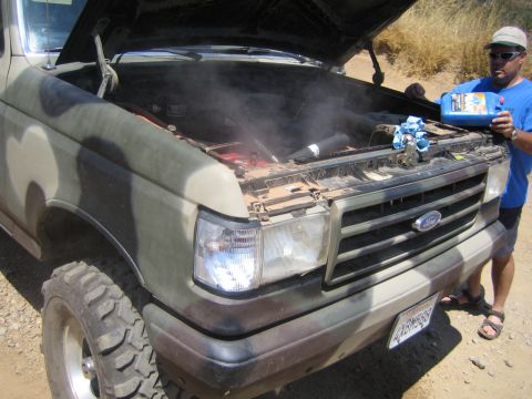 1990 Ford Bronco - man in a blue shirt pours coolant into the steaming engine bay of a camouflage-painted Ford truck with the hood raised.