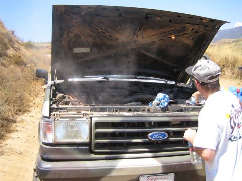 1990 Ford Bronco - man holding water bottles inspects the steaming engine bay of a Ford truck with its hood raised on a dirt trail.
