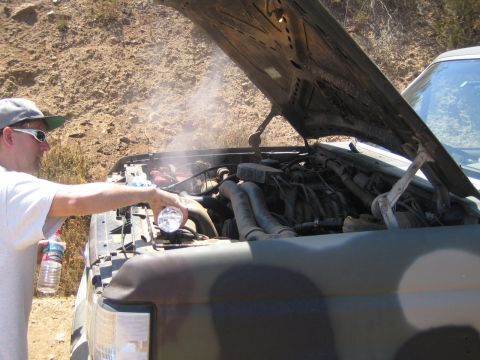 1990 Ford Bronco - man in sunglasses reaches into the steaming engine bay of a camouflage-painted Ford truck with the hood raised on a trail.