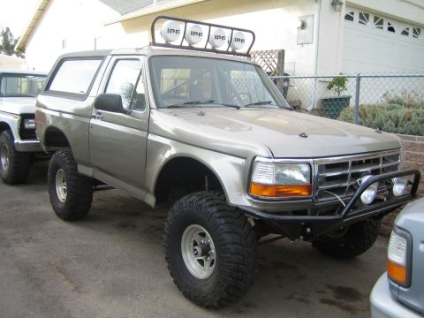 1990 Ford Bronco - tan lifted Ford Bronco with a four-light roof bar and tube front bumper parked in a driveway beside a house.
