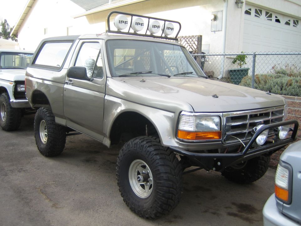 Tan lifted Ford Bronco with roof light bar and tube bumper parked in a residential driveway
