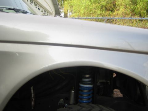 1990 Ford Bronco - Close-up of a silver fender with the wheel removed, revealing a blue-and-white coilover shock in the wheel well.