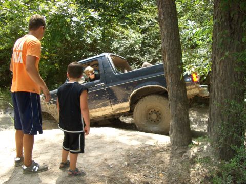 1990 Ford Bronco - A Ford Bronco is shown off-roading on a dirt trail surrounded by trees, with two onlookers observing its progress.