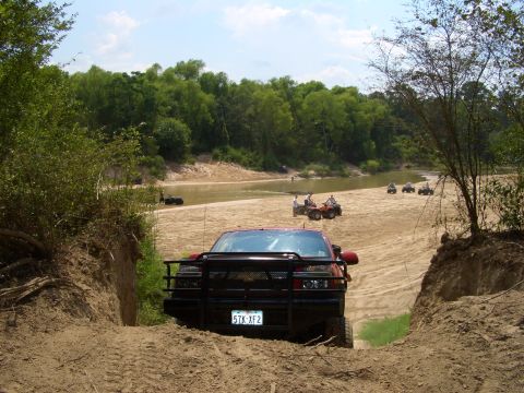 1990 Ford Bronco - Several ATVs are parked and being ridden near a riverbank while a red pickup truck navigates a sandy trail.