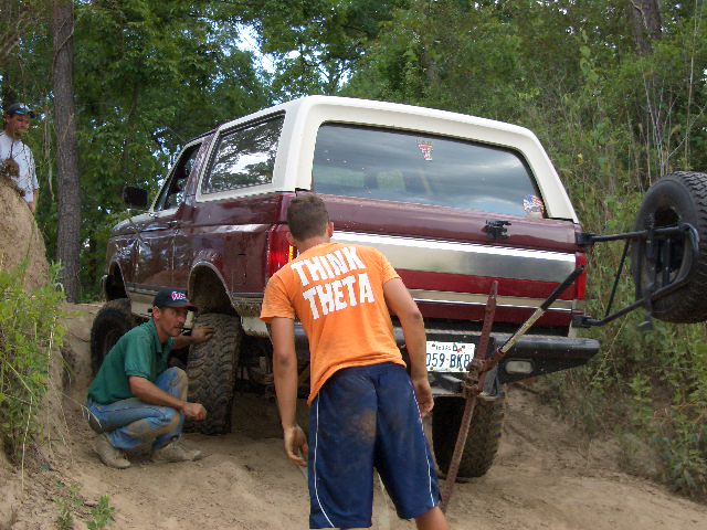 Maroon Ford Bronco with white top on a dirt trail, people working on the rear tire