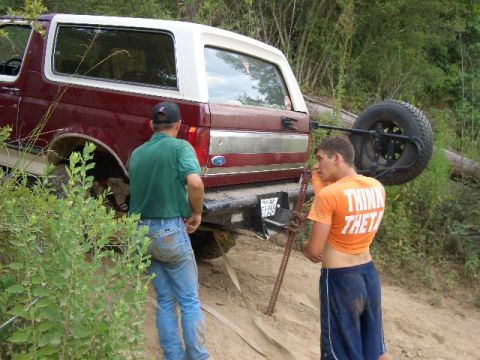 1990 Ford Bronco - Two men attempt to free a maroon and white Ford Bronco stuck in the dirt, with a spare tire mounted on the rear.