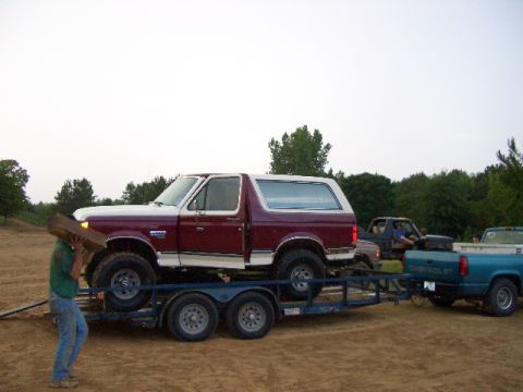 1990 Ford Bronco - A person carries wood while a Ford Bronco is loaded onto a trailer, with other trucks in the background.
